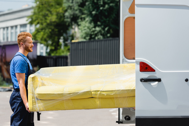 Vista lateral del cargador en sofá de sujeción uniforme en abrigo elástico cerca de camión con puertas abiertas en la calle urbana  - Foto, Imagen