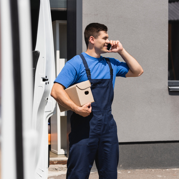 Selective focus of loader talking on smartphone and holding cardboard box near truck on urban street  - Fotografie, Obrázek