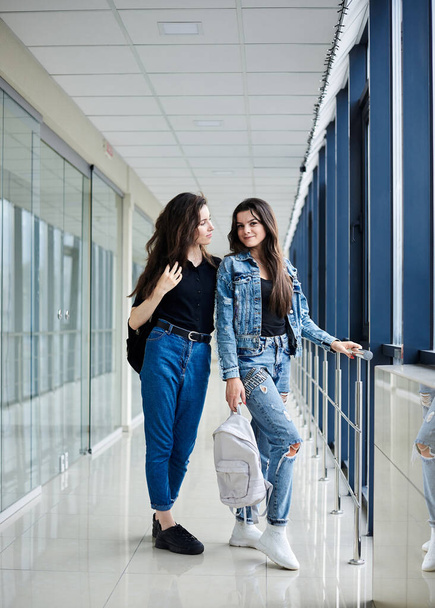 Two young brunette girls, standing in light airport hallway with huge windows,wearing casual jeans clothes, holding white and black backpacks.Girlfriends, waiting for flight, smiling. Safe air travel. - Photo, Image