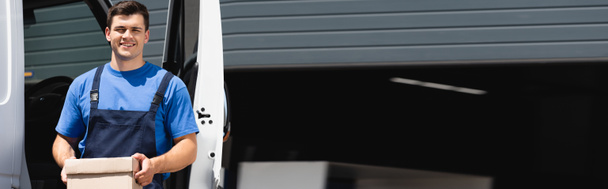 Vízszintes kép a rakodóról, ahogy a kamerát nézi és kartondobozt tart a teherautó mellett a szabadban  - Fotó, kép