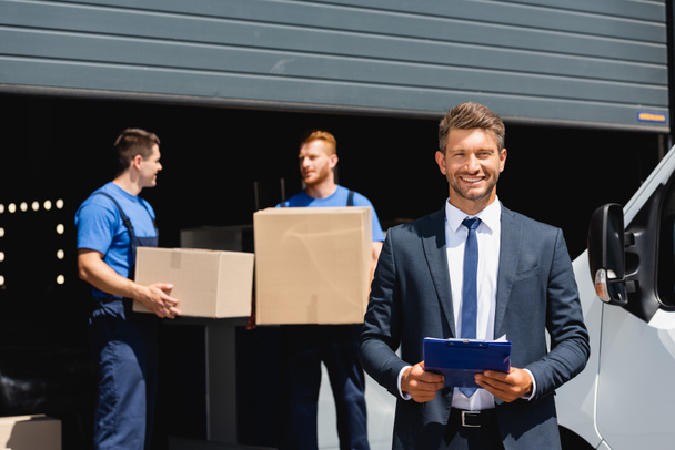 Concentration sélective de l'homme d'affaires tenant presse-papiers tandis que les déménageurs tenant des boîtes en carton près du camion et de l'entrepôt  - Photo, image
