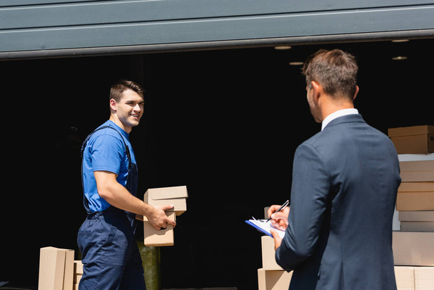 Concentration sélective de chargeur tenant des boîtes en carton près de l'homme d'affaires écrivant sur le presse-papiers et l'entrepôt à l'extérieur - Photo, image