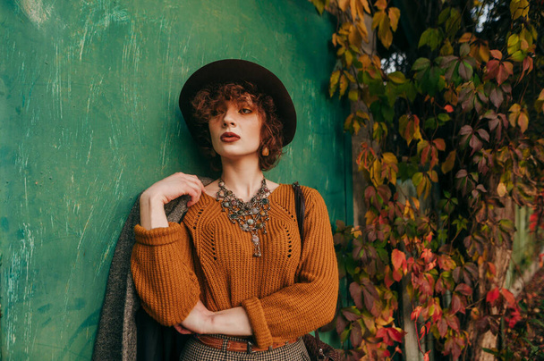 Fashionable lady in vintage clothing stands on background of green grunge wall and yellowed autumn ivy and looks side with a serious face. Model curly girl posing in the city on a rustic background. - Foto, imagen