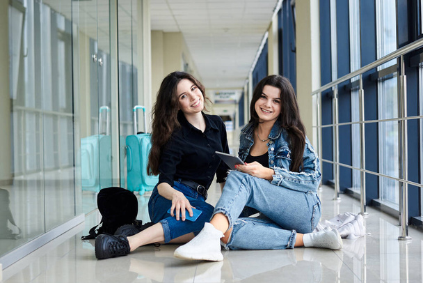 Two young brunette girls, sitting on floor in light airport hallway, with luggage behind, checking phone, smiling, wearing casual jeans clothes. Girlfriends, traveling by air, waiting for flight. - Photo, Image