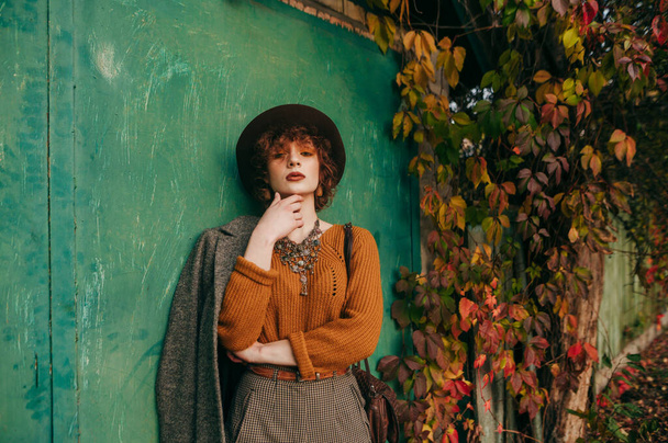 Stijlvolle krullende dame in vintage kleding en hoed poseert voor camera met serieus gezicht tegen groene grunge muur en oranje klimop. Herfst portret van modieuze dame op het platteland. - Foto, afbeelding