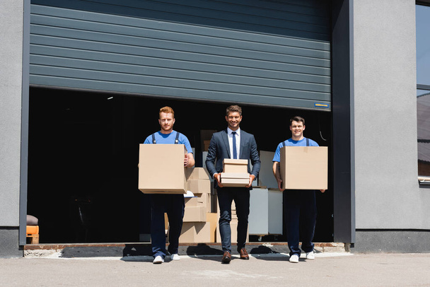 Businessman in suit and movers looking at camera while holding carton boxes near warehouse on urban street  - Photo, Image