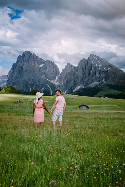para mężczyzn i kobiet na wakacjach w Dolomitach Włochy, Alpe di Siusi - Seiser Alm z grupą górską Sassolungo - Langkofel w tle o zachodzie słońca. Żółte wiosenne kwiaty i drewniane domki w - Zdjęcie, obraz