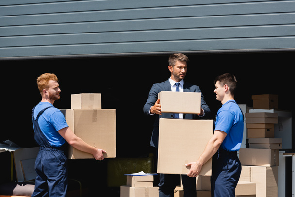 businessman in formal wear stacking packages near movers working outdoors - Fotografie, Obrázek