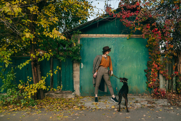 Modna pani pozująca z psem na tle zielonej ściany wiejskiego domku i drzew z żółtymi liśćmi, trzymająca zwierzaka na smyczy. Jesienne zdjęcie pięknej dziewczyny z psem w wiosce - Zdjęcie, obraz