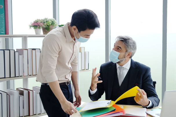 シニア白人の上司と若いビジネスマンは、コヴィド19ウイルスが文書シートを保持し、オフィスでの仕事について話すのを防ぐマスクを着用。社会的距離と新しい通常のライフスタイルコンセプト - 写真・画像