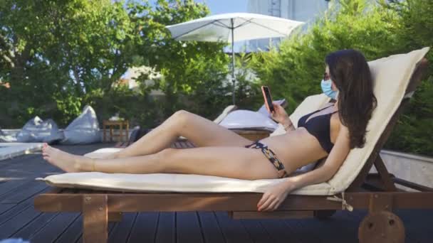 Mujer en bikini tumbada en una tumbona de madera en la piscina, revisando las noticias en su teléfono inteligente. Mujer con mascarilla en la piscina - Metraje, vídeo