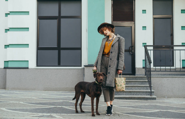 Fashionable portrait of a stylish lady in a coat and hat on a walk with a pet on the street of the town, standing against a wall and holding a leash with dog in hand. Girl stands with dog on building - Foto, Bild