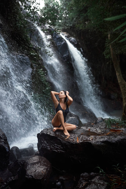 beautiful brunette sits on a stone in the background of a waterfall washing her face under i - Foto, Imagem