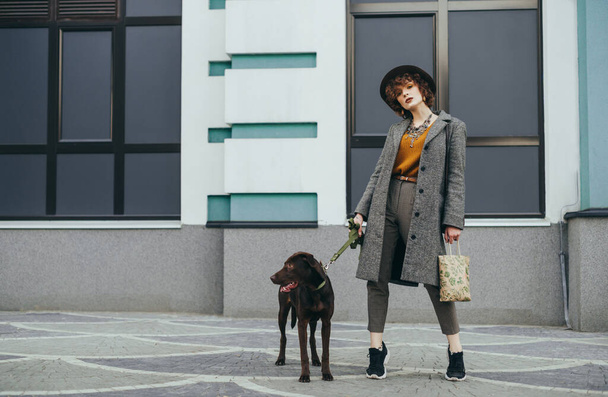 Divatos hölgy kabátban és kalapban áll kutya pórázon fal háttér, belenéz a kamerába komoly arc és a csomagolás a kezében. Divatos fotó egy nőről kutyával az utcán.. - Fotó, kép