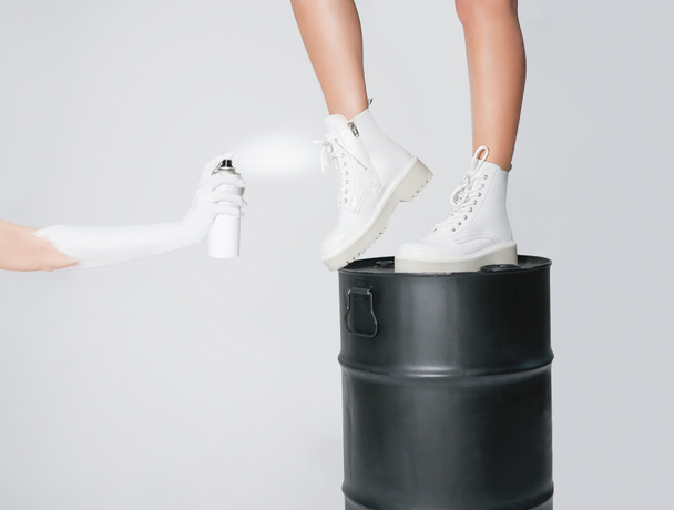                   the girl takes care of white leather shoes by painting boots with an aerosol spray. woman legs in fashionable white shoes on a light background. close-up                                            - Foto, Imagen