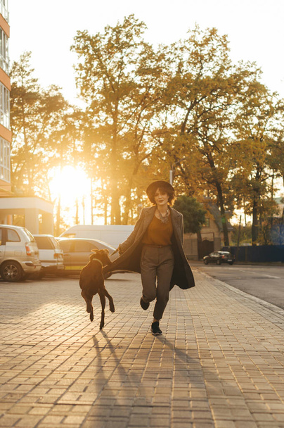 Κομψό κυρία με καπέλο και παλτό τρέχει γύρω με παιχνιδιάρικο σκυλί σε ένα λουρί στο φόντο των δέντρων του φθινοπώρου στο ηλιοβασίλεμα. Βραδινή βόλτα το ηλιοβασίλεμα με ένα κατοικίδιο. Κάθετη - Φωτογραφία, εικόνα