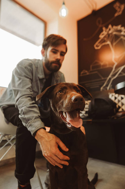 Bärtiger Mann sitzt in gemütlichem Café mit Hund. Porträt eines Hundes, der drinnen mit einem Haustier spielt. Vertikal - Foto, Bild