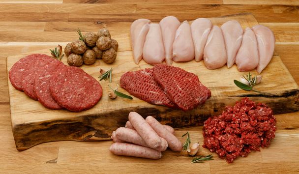 Eine Platte mit rohem Fleisch, inkl. Rind, Huhn und Schwein, mit Knoblauch und Kräutern auf einem Holzbrett, auf einer Holzbank. - Foto, Bild