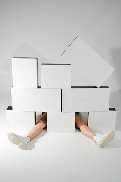 una chica se sienta en zapatos blancos sobre un fondo claro entre grandes cajas en blanco y negro. zapatos embalaje publicidad zapatos de moda                        - Foto, Imagen