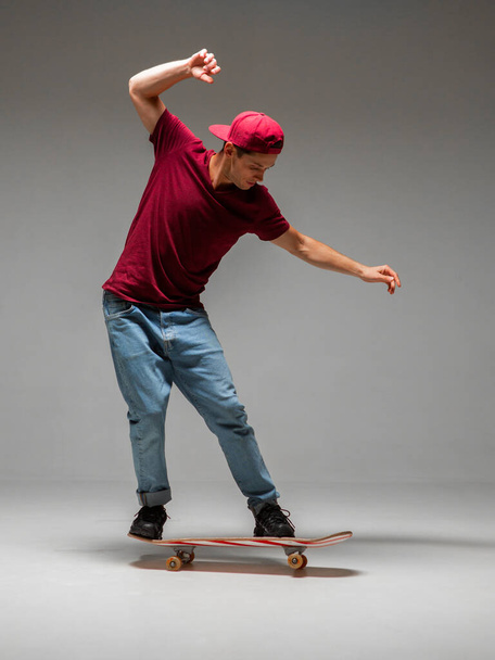 Cool young guy skateboarder in red baseball cap rides on a skateboard in studio on grey background - Foto, immagini