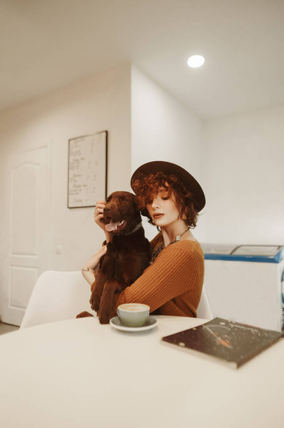 Retrato de una chica con estilo en un sombrero se sienta en un acogedor café, acariciar a un perro y beber café. La dama con una mascota en sus brazos pasa tiempo en un café amigable con los animales. Vertical. - Foto, imagen