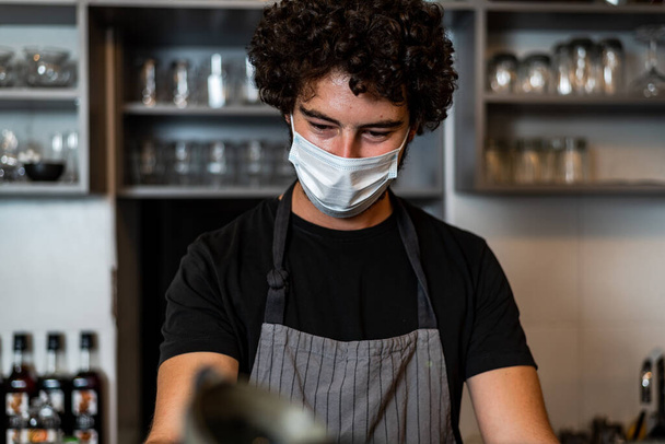 Молодой человек, бариста, делает кофе эспрессо во время ношения хирургической маски и перчаток для предотвращения распространения коронного вируса - Концепция работы бара по безопасности. - Фото, изображение