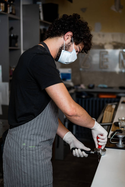 Junger Mann, Barista, macht Kaffee-Espresso und trägt dabei Chirurgenmaske und Handschuhe zur Verhinderung der Ausbreitung des Coronavirus - Sicherheitskonzept der Bar. - Foto, Bild