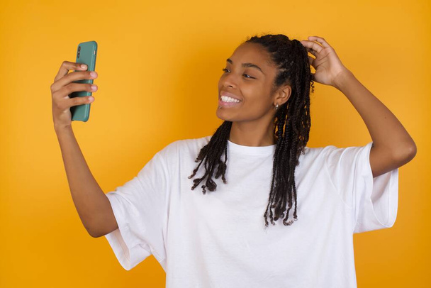 Fotografie okouzlující mladá žena tmavé pleti s copánky vlasy na sobě ležérní oblečení s úsměvem a přičemž selfie připraven k odeslání na její sociální média. - Fotografie, Obrázek