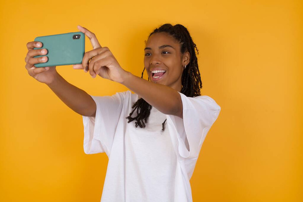 Portret z bliska Młoda ciemnoskóra kobieta z warkoczami, ubrana w luźne ubrania, robiąca selfie, aby zamieścić je w mediach społecznościowych lub wykonująca wideo rozmowę z przyjaciółmi. - Zdjęcie, obraz