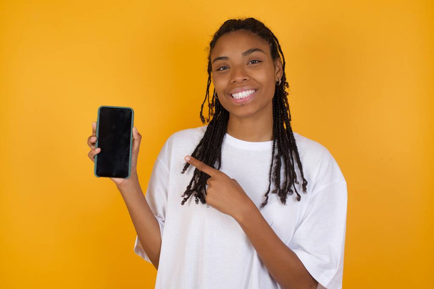 cheerful attractive Young dark skinned woman with braids hair wearing casual clothes with telephone in hand new model of apple telephone just have look at price - Foto, Bild