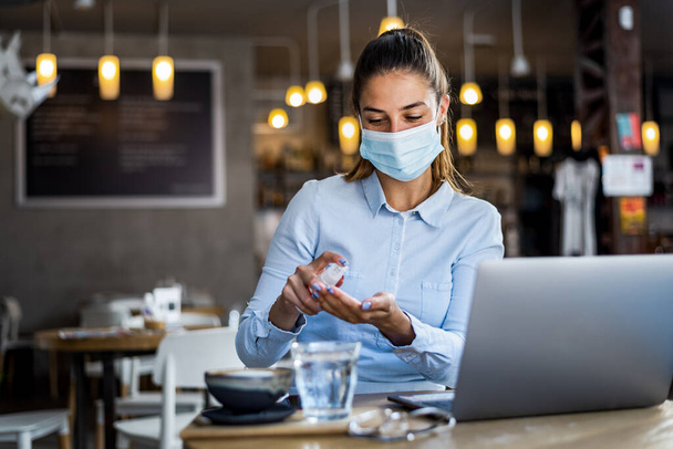 Портрет молодой женщины в хирургической маске с использованием геля для дезинфекции рук в ресторане. Antiseptic, Pandemic Corona virus, Hygiene and Healthcare concept - Фото, изображение