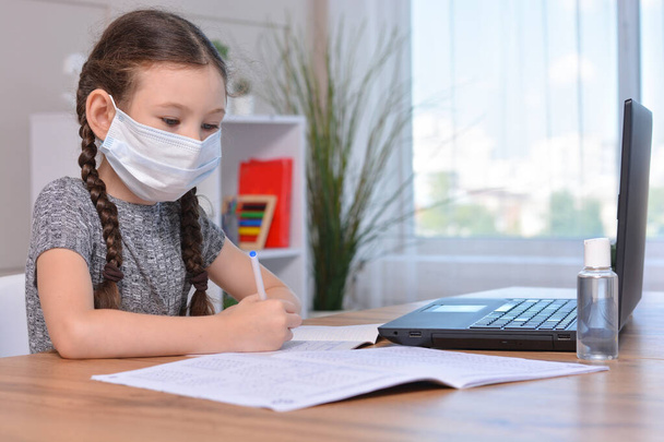 A schoolgirl in a medical mask is doing her homework on a laptop at home or studying in class at a lesson at school.Student safety after covid-19 pandemic. Back to school.Distance learning. - Zdjęcie, obraz