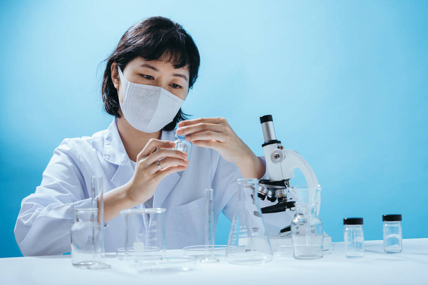 Intelligente giovane scienziata asiatica in camice da laboratorio con maschera medica. Ispezione sugli strumenti di laboratorio. Spazio vuoto in studio girato isolato su sfondo blu colorato. Concetto medico. - Foto, immagini