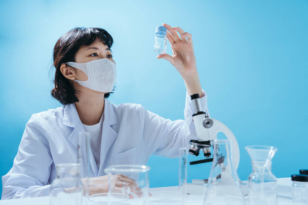 Розумна молода азіатська жінка-вчена в лабораторному пальто з медичною маскою для обличчя. Інспекція на лабораторних інструментах. Порожній простір у студійному знімку ізольовано на барвистому синьому фоні. Медична концепція
. - Фото, зображення