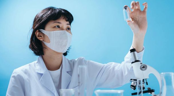 研究室のコートのスマート若いアジアの女性科学者医療面マスク。実験器具の検査。スタジオでの空のスペースは、カラフルな青の背景に隔離されたショット。医学的概念. - 写真・画像