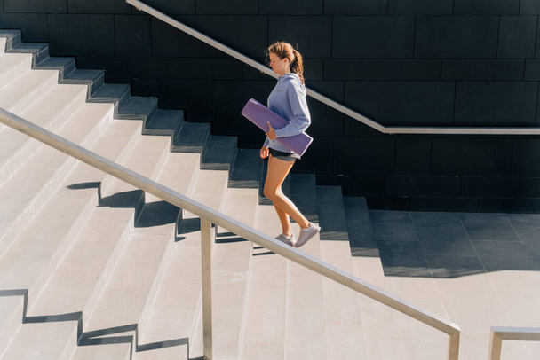 Stylish young girl in sportswear runs on the steps among the skyscrapers in the fresh air for weight loss. Summer sunset fitness workout concept, athletics for weight loss - Photo, Image