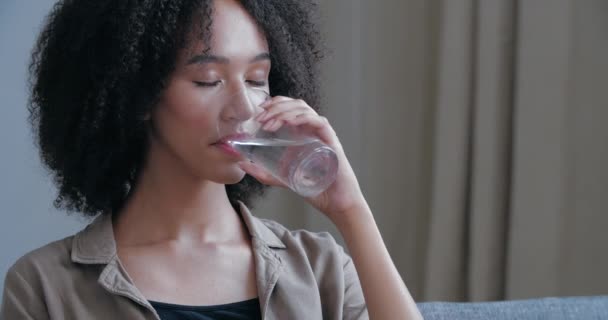 Közelkép fiatal afro-amerikai nő oltja szomját, tartja üveg hideg meleg víz, iszik, becsukja a szemét öröm, mosolygós néz a kamerába. Vonzó göndör hajú lány frissítő - Felvétel, videó