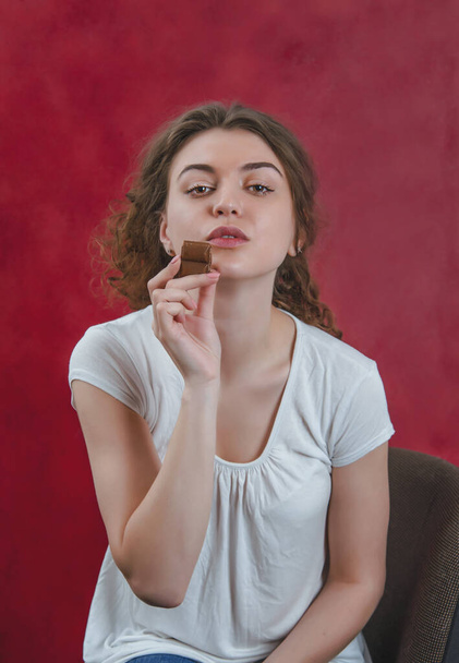 Pretty girl eating chocolate. Portrait on red background, delicious food, sweet chocolate. - Photo, image
