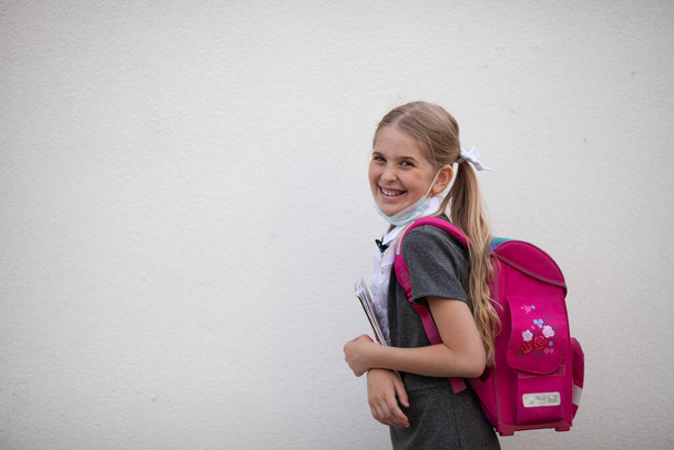 Счастливая школьница снимает медицинскую маску с лица и улыбается. Блондинка с розовым рюкзаком держит книгу в руках. Счастливые дети идут в школу после карантина. Фон белой стены с пробелами для текста.  - Фото, изображение