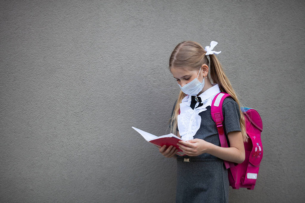 Μια μαθήτρια που φοράει ιατρική μάσκα και κουβαλάει ένα ροζ σακίδιο μένει κοντά στο σχολείο της, κοιτάζει στο βιβλίο εργασίας, επαναλαμβάνει τα μαθήματά της. Πίσω στο σχολείο. Γκρι φόντο τοίχου με χώρο για κείμενο. - Φωτογραφία, εικόνα