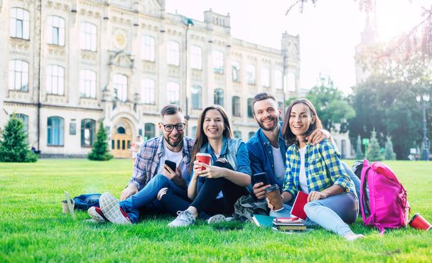 Happy beautiful students team with backpacks and other stuff are talking, having fun and relaxing on the grass on college background outdoors - 写真・画像