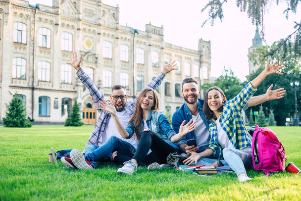 Happy beautiful students team with backpacks and other stuff are talking, having fun and relaxing on the grass on college background outdoors - 写真・画像