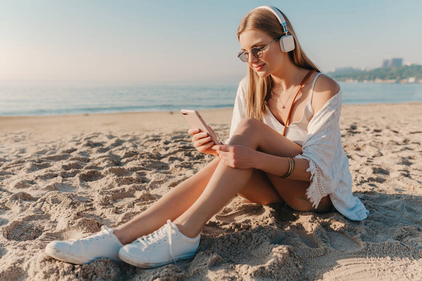 stylish young attractive blond smiling woman sitting on beach in headphones listening to music relaxed - Foto, afbeelding