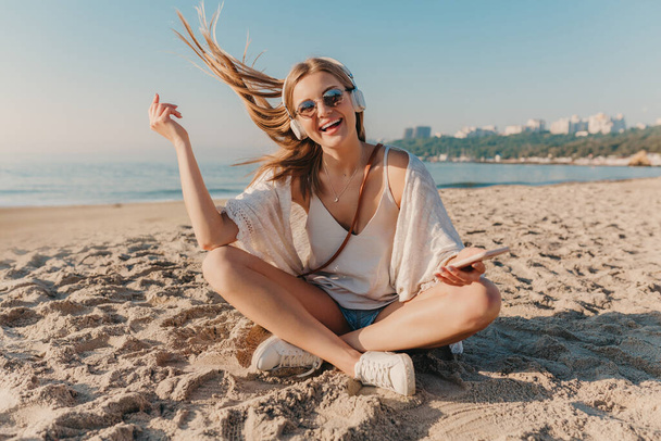 young attractive blond smiling woman sitting on beach in headphones listening to music in positive happy mood - Photo, Image