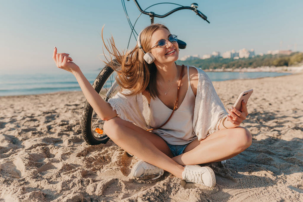 stylish young attractive blond smiling woman sitting on beach with bicycle in headphones listening to music relaxed using phone positive mood - Foto, Imagen