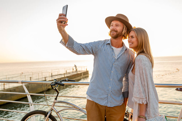 young attractive smiling happy man and woman traveling on bicycles taking selfie photo on phone camera, romantic couple by the sea on sunset, boho hipster style outfit, friends having fun together - Foto, Bild