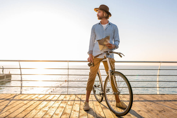 young attractive man traveling on bicycle by sea on summer vacation by the sea on sunset, boho hipster style outfit, holding map sightseeing with camera, dressed in shirt and hat - Фото, изображение