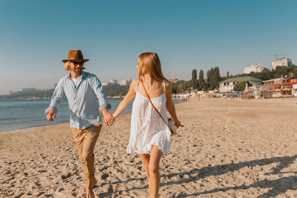 young attractive smiling happy man in hat and blond woman running together on beach on summer vacation traveling, romantic couple by the sea on sunset, boho hipster style outfit wearing sunglasses, friends having fun together - Photo, Image