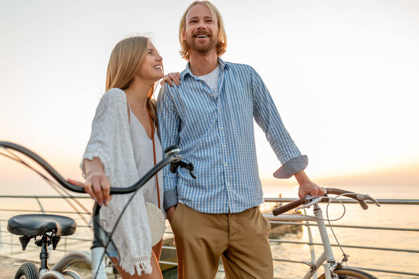 attractive happy couple embracing in love traveling in summer on bicycles, man and woman with blond hair boho hipster style fashion having fun together, walking by the sea in resort city - Valokuva, kuva