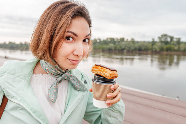 Asiatisches glückliches Mädchen isst ein frisches französisches Dessert Eclair und trinkt Kaffee. Street Food zum Mitnehmen. Freizeit und Erholung am Stadtdamm - Foto, Bild